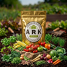 A gold resealable pouch labeled ARK Backyard Kit stands in a garden bed surrounded by fresh vegetables including tomatoes, corn, beans, carrots, broccoli, and herbs.
