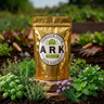 A gold resealable pouch labeled ARK Herbdom Seeds Herb Seed Kit stands on a stone in a garden bed surrounded by green herbs and soil.