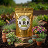 A gold resealable pouch labeled ARK Heirloom Seeds Patio Seed Kit stands on a stone in a garden setting, surrounded by small potted plants and flowers.
